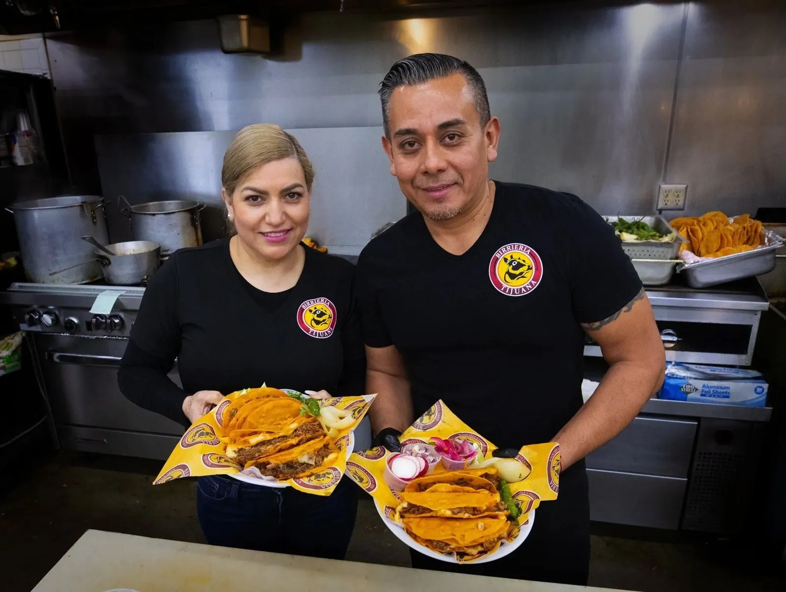 Jenny and Freddy, owners of Birrieria Tijuana, holding plates of tacos